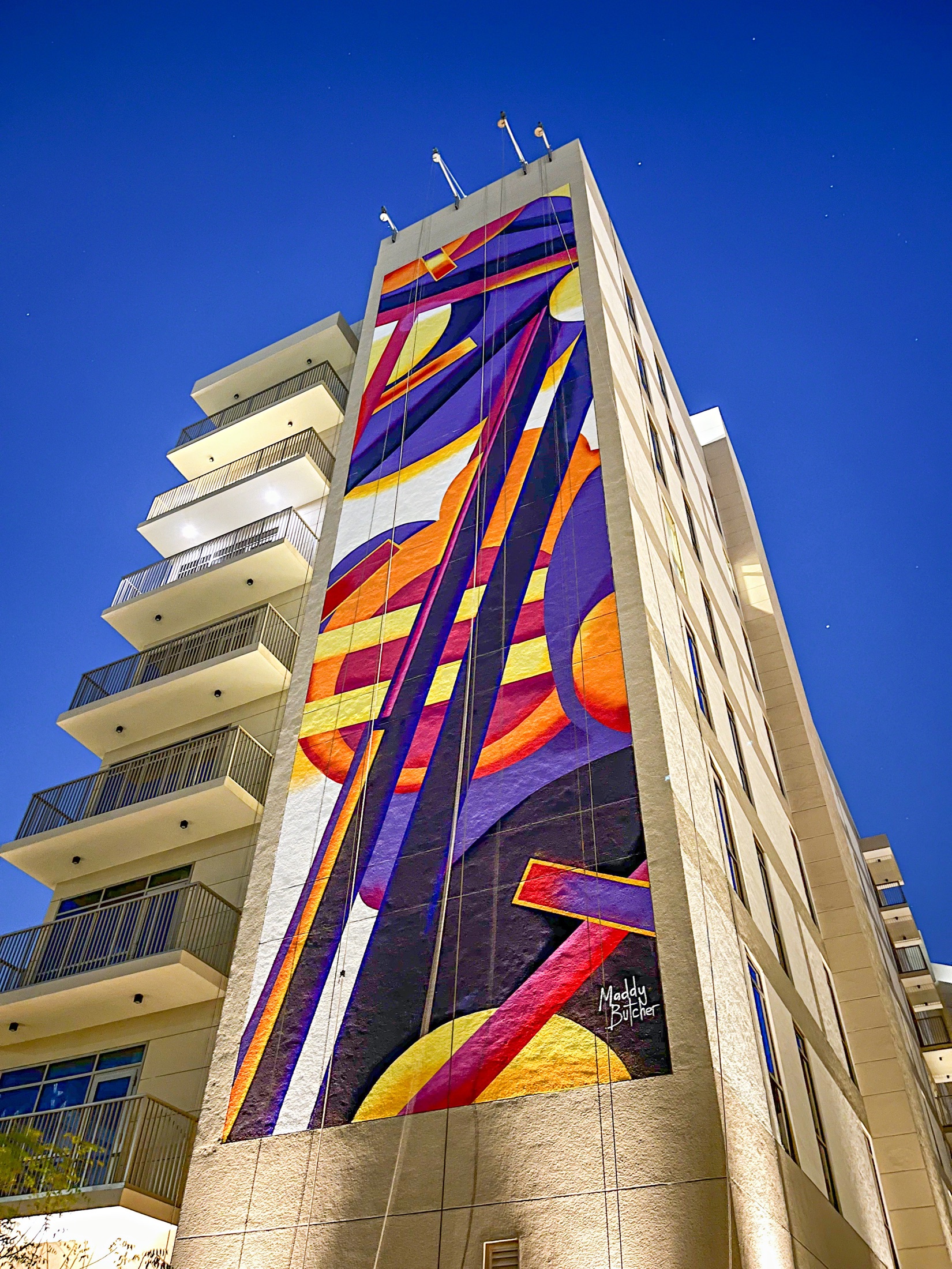 Maddy Butcher’s completed purple and orange abstract mural rising across an Aldar building at night